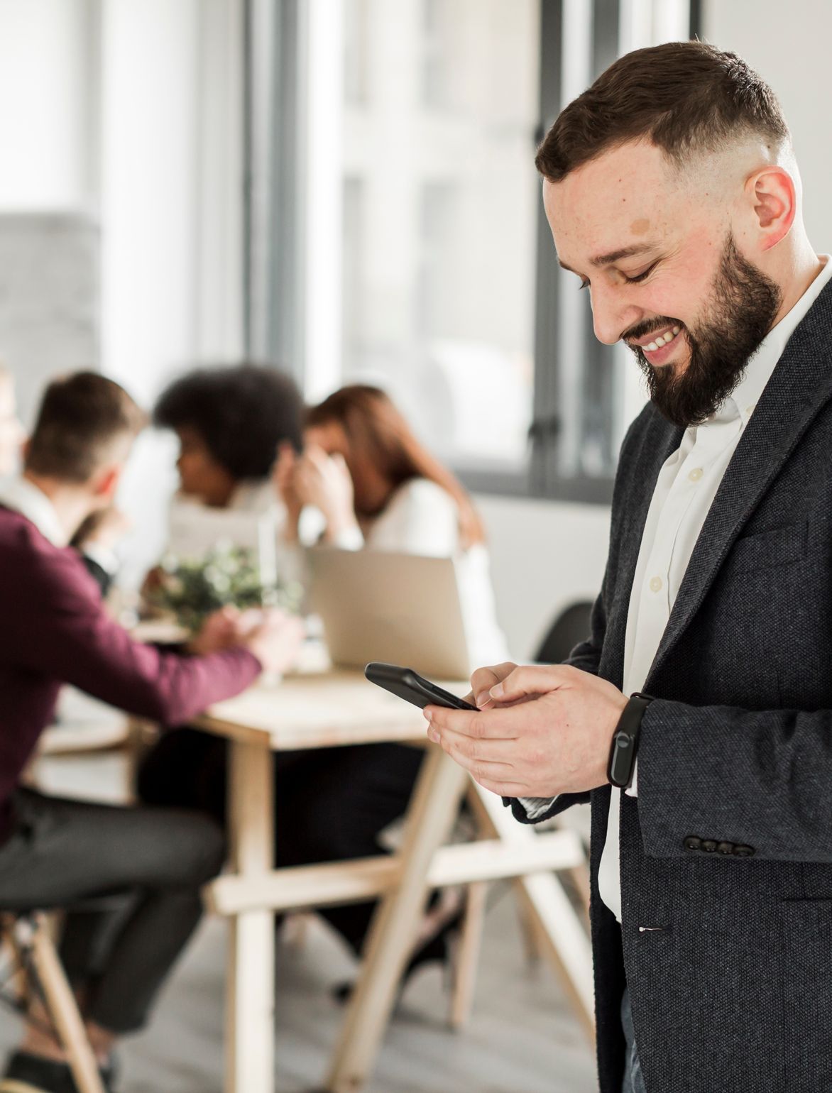 A man smiling at his phone while a meeting is taking place in the background